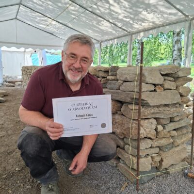 Účastník kurzu drží certifikát u postavené kamenné zdi