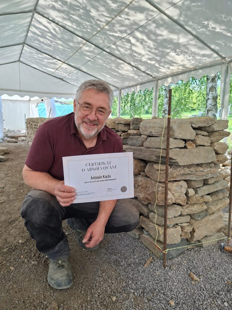 Účastník kurzu drží certifikát u postavené kamenné zdi